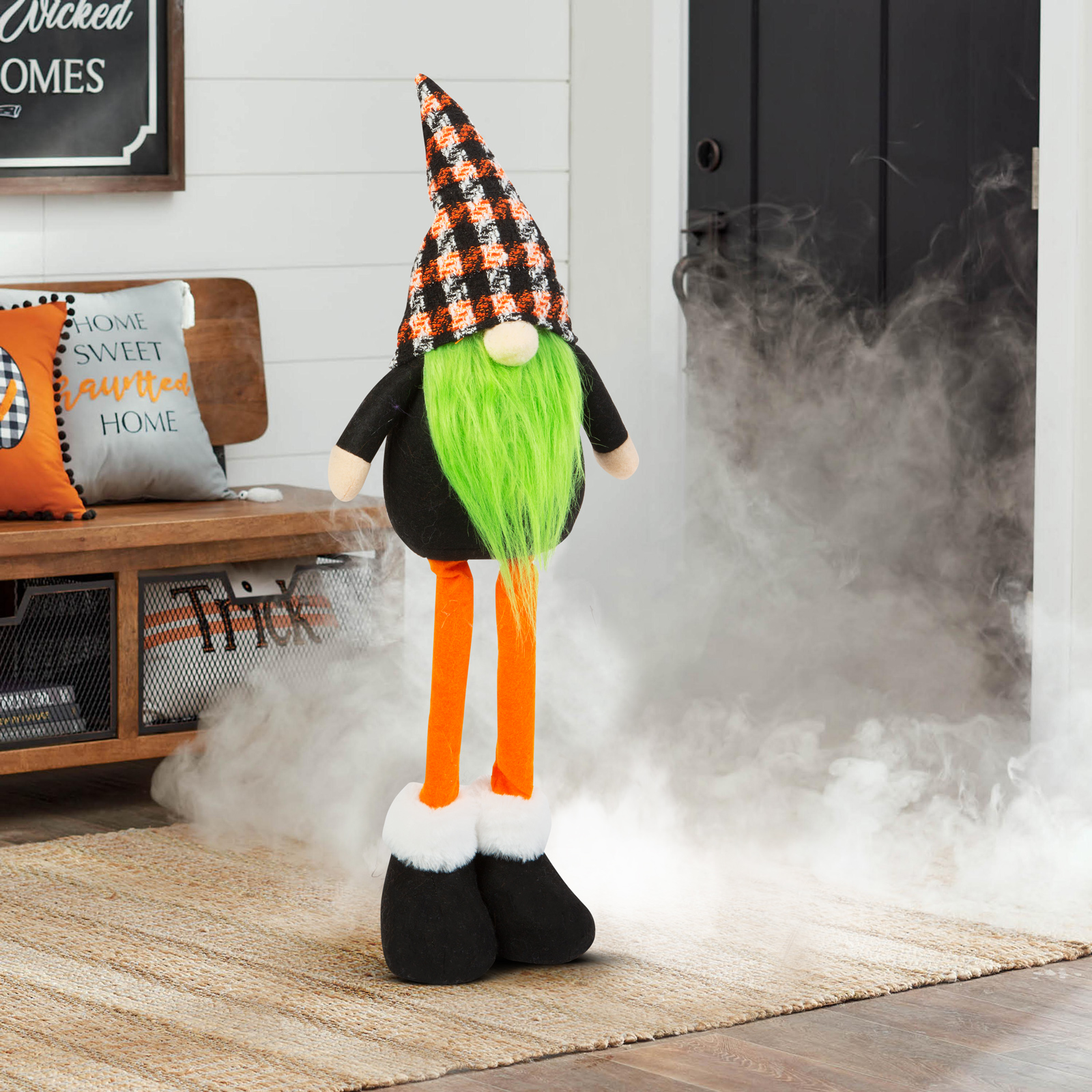 Halloween-i gnóm hosszú narancssárga lábakkal, zöld szakállal és kockás kalappal, bent állva.