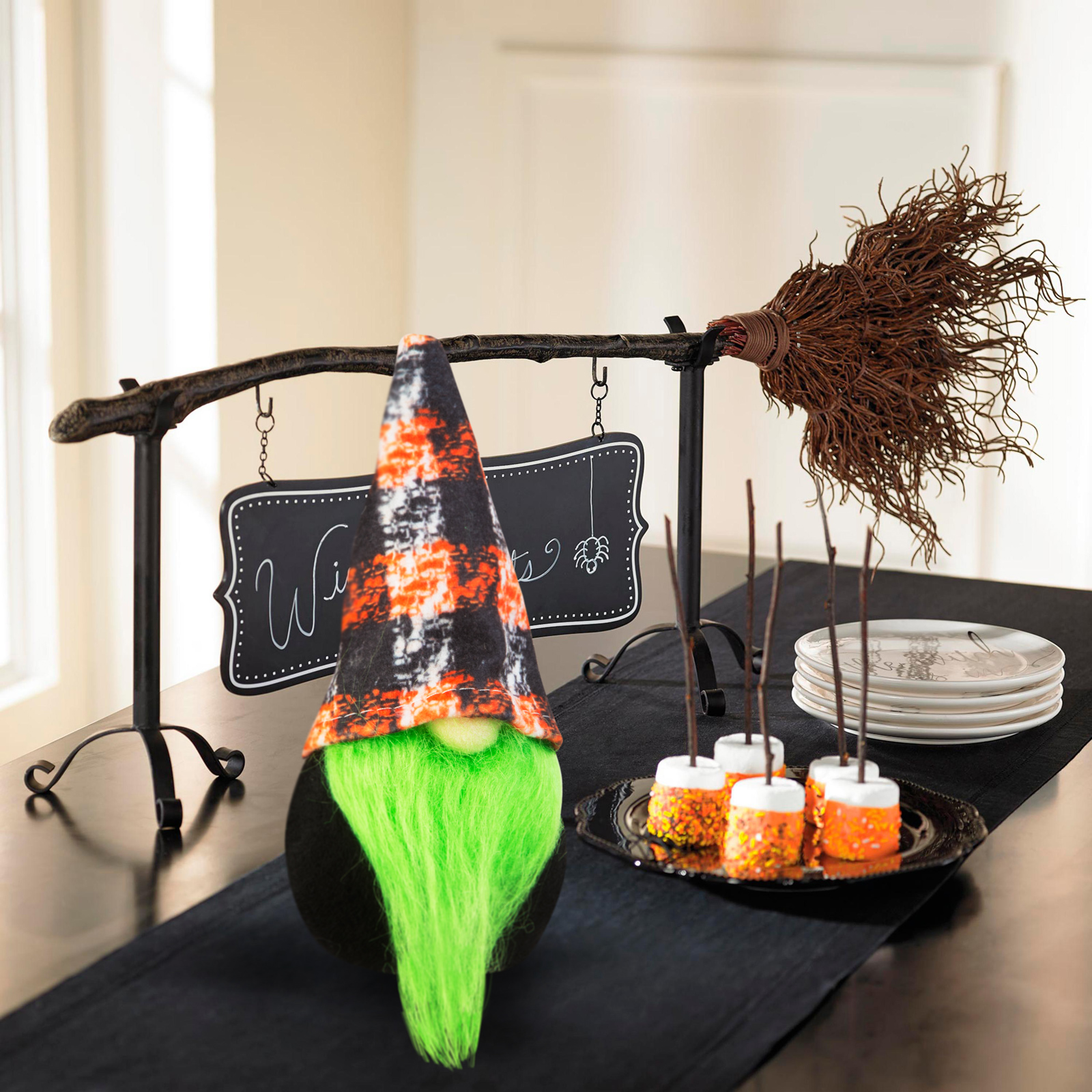 Halloween-gnóm narancs-fekete kalappal, zöld szakállal, seprűvel, táblával és édességekkel a fekete asztalon.