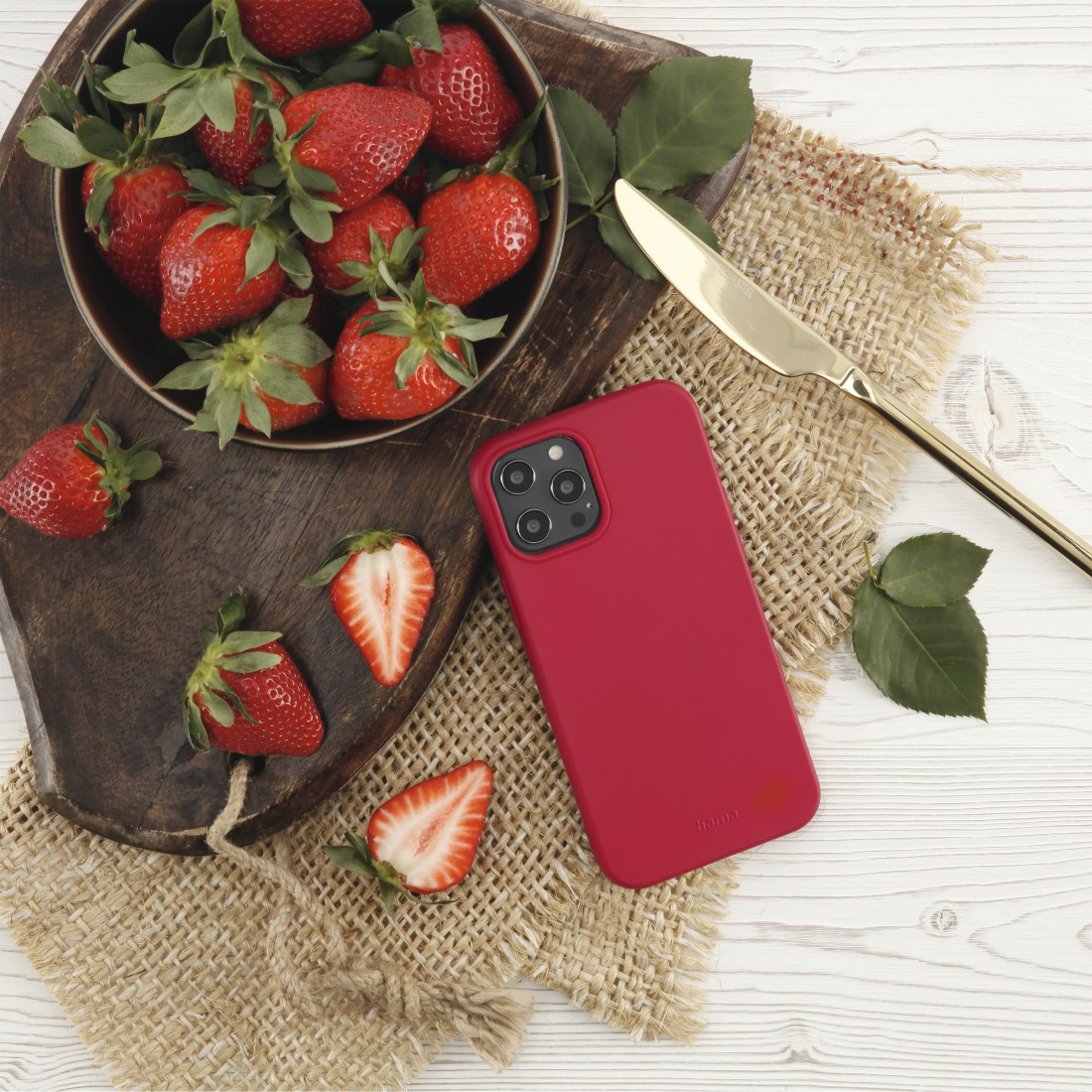Eine Schüssel Erdbeeren, eine rote Handyhülle, ein Messer und einige Blätter auf einem Holzbrett und einem Leinentuch.
