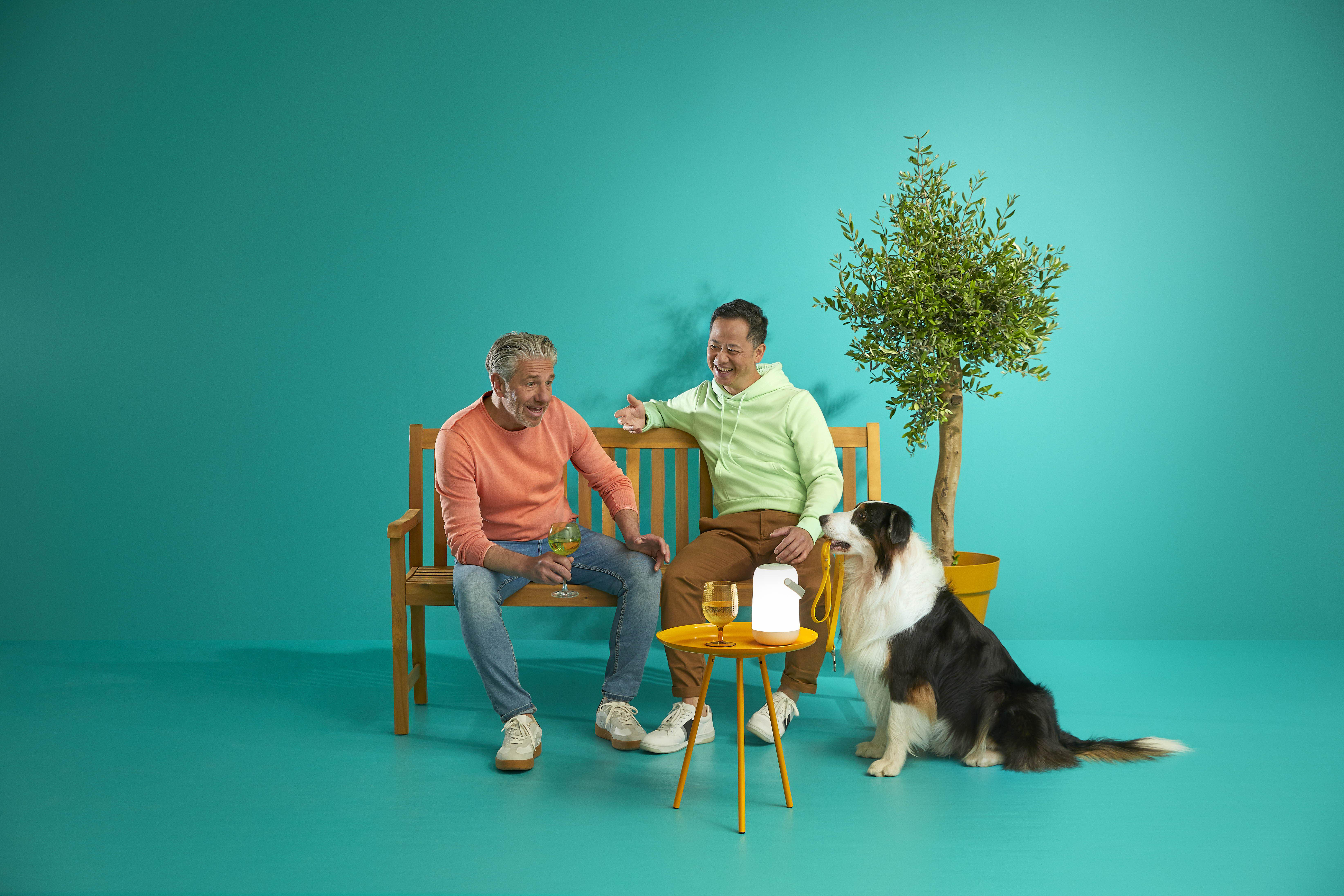 Zwei Männer und ein Hund sitzen draußen auf einer Bank mit Getränken und einer Lampe.