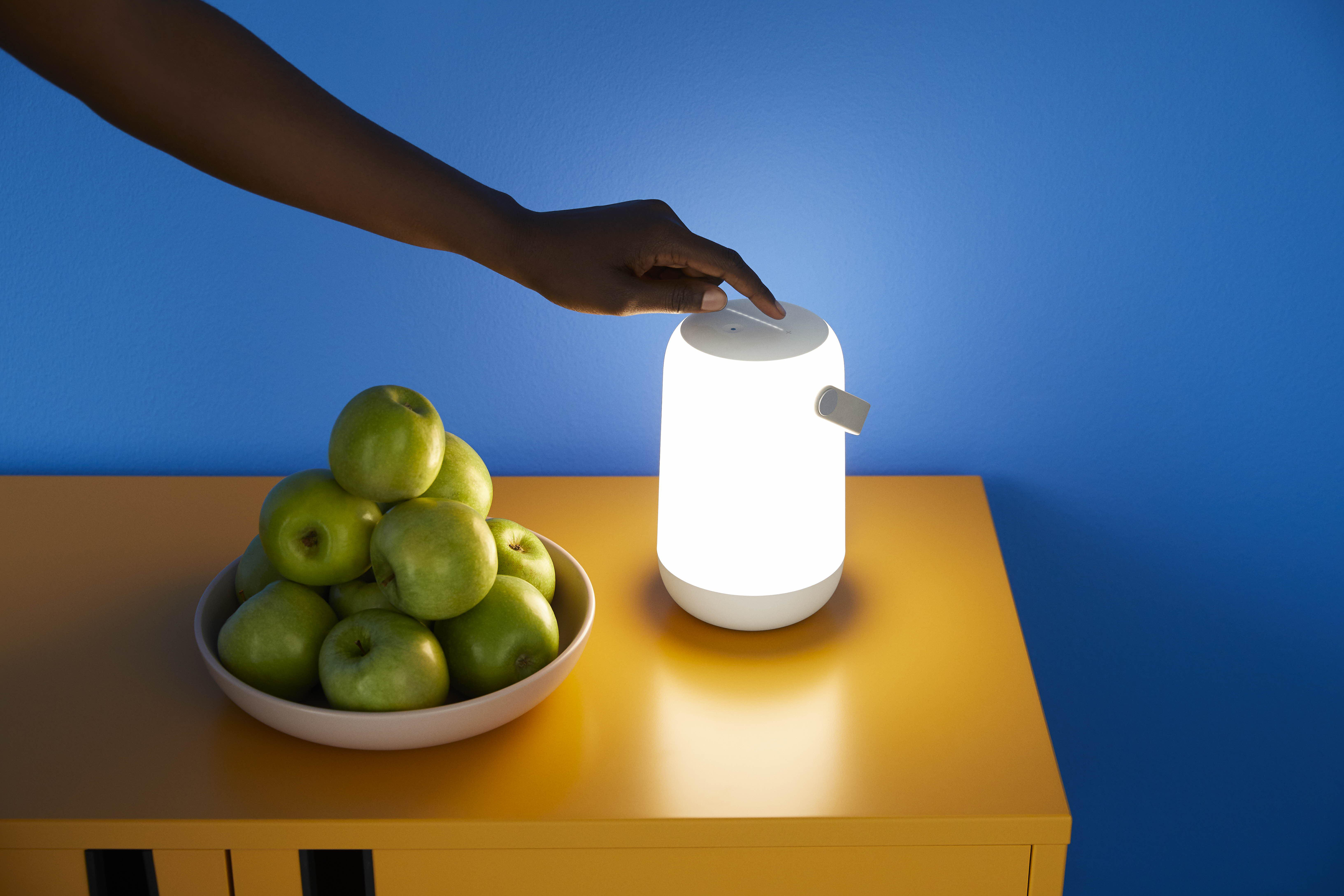 Hand schaltet weiße Lampe auf gelbem Tisch an, grüne Äpfel in Schale, blauer Hintergrund.