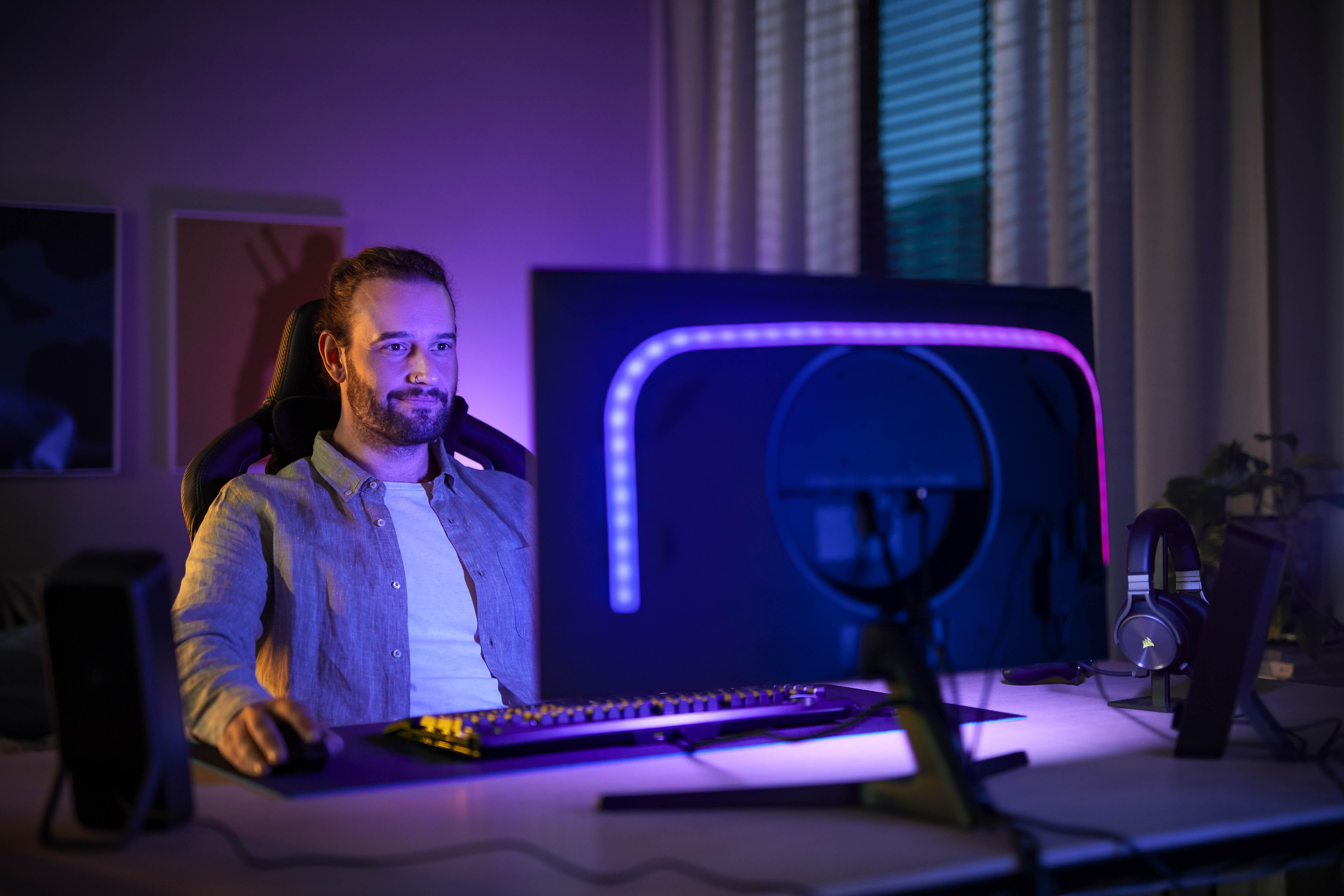 Ein Mann in einem Gaming-Setup mit Monitor, Tastatur und Kopfhörern auf einem dunklen Schreibtisch.