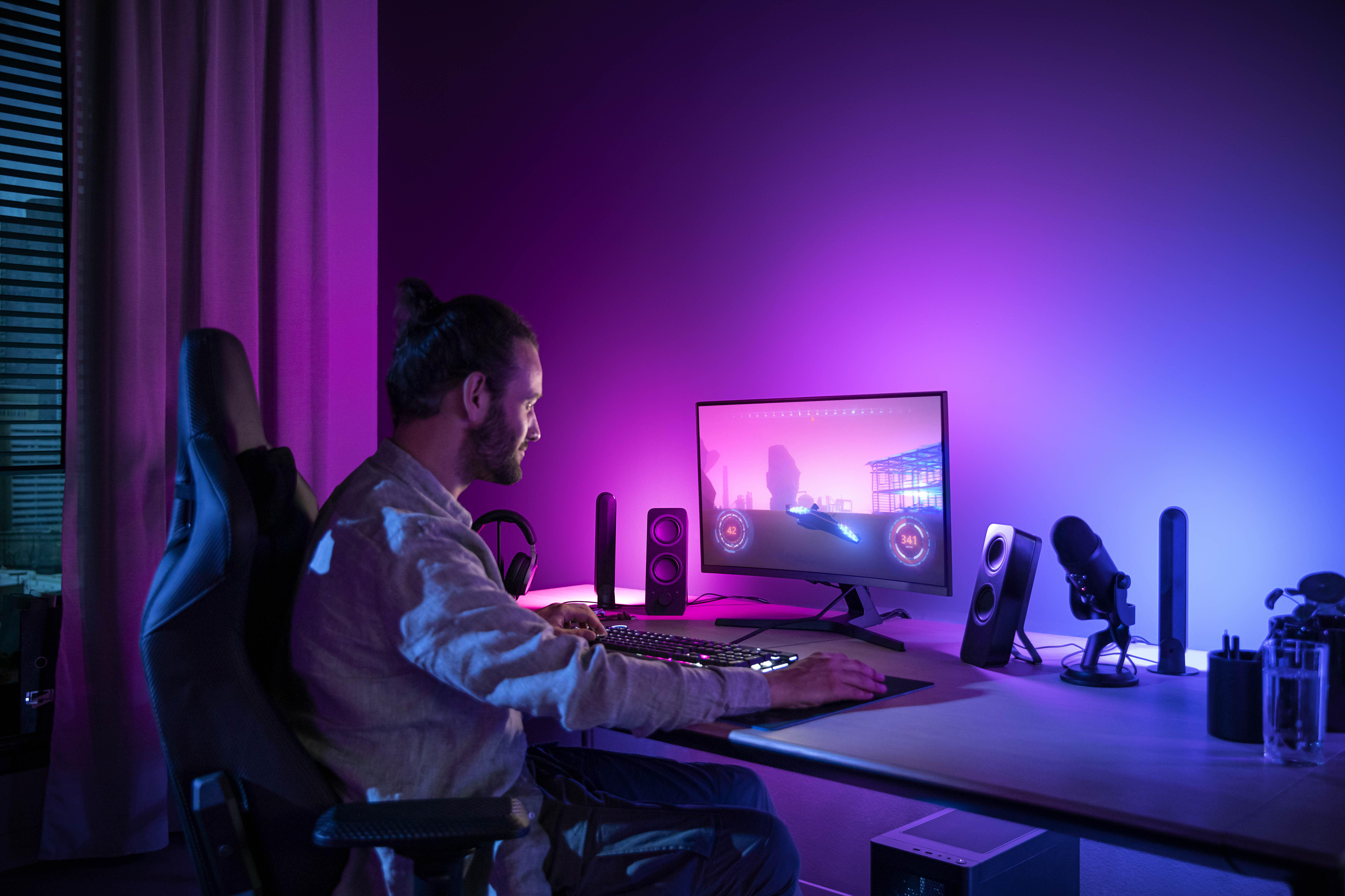 Mann, der am Schreibtisch spielt. Monitor, Tastatur, Lautsprecher, Mikrofon, Headset und lila Beleuchtung.