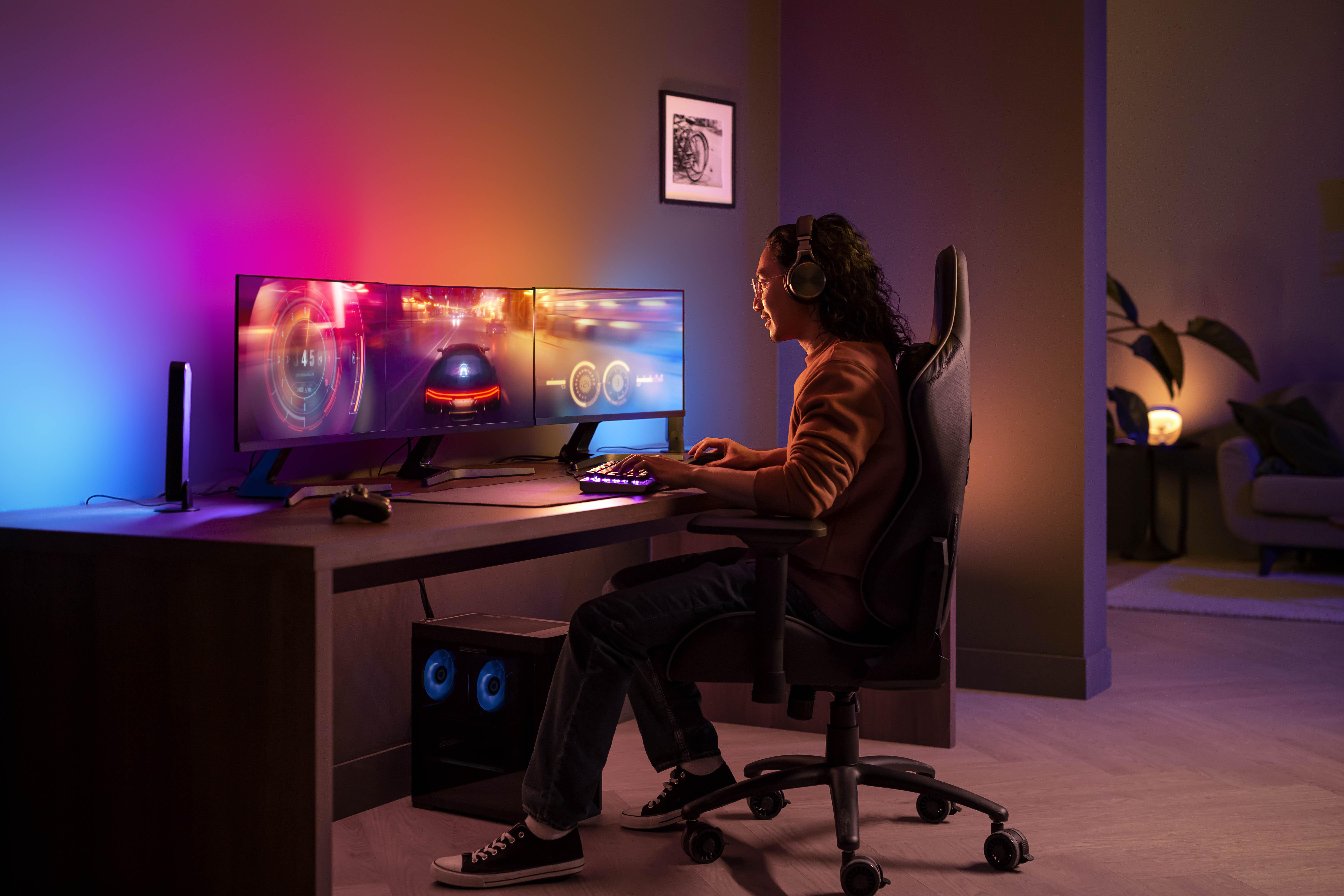 Eine Person spielt mit einem Dreifach-Monitor-Setup, verwendet eine Tastatur mit farbenfroher Ambientebeleuchtung.