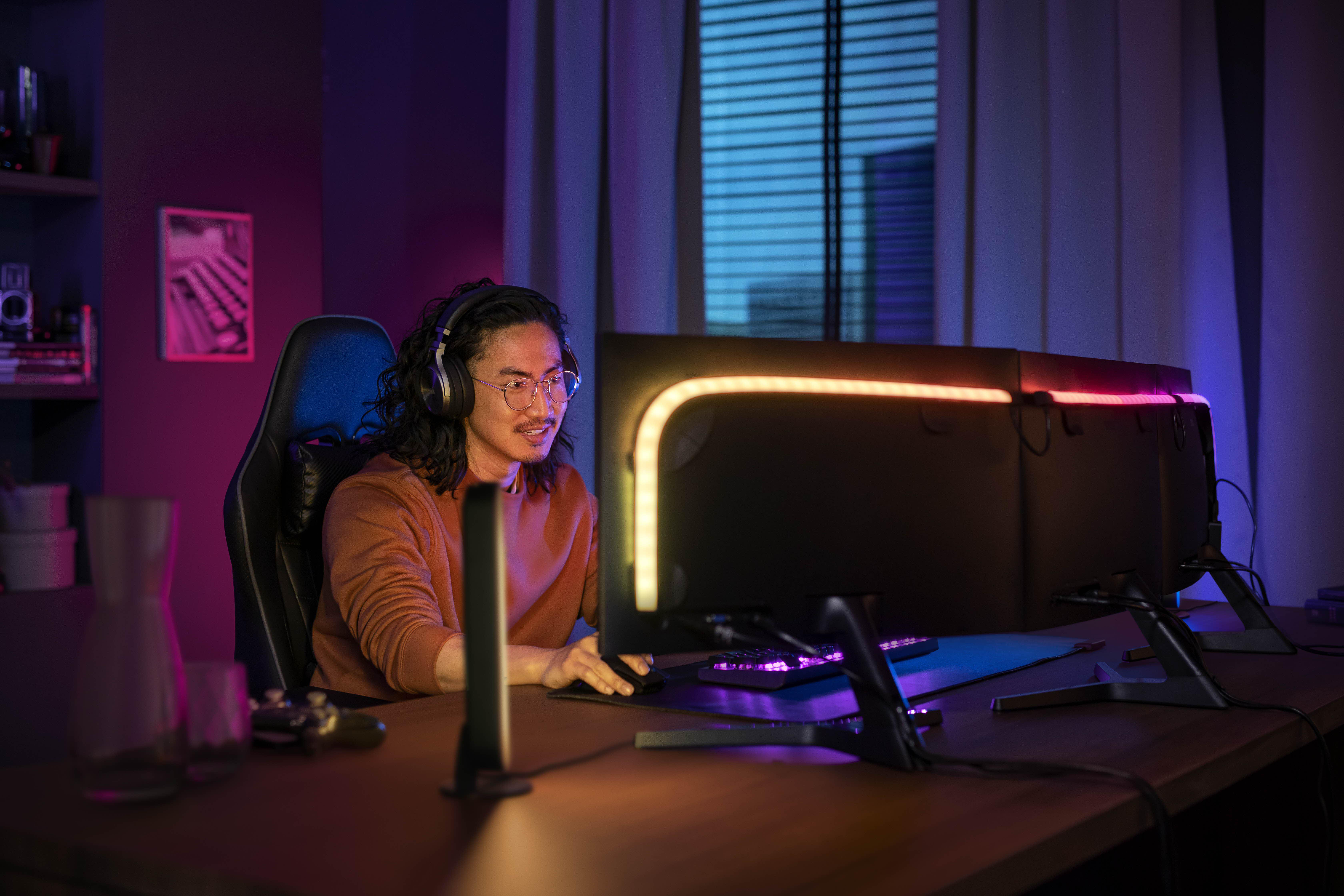 Ein Mann, der nachts spielt. Er trägt ein Headset. Auf dem Schreibtisch sind Monitore und Lichter.