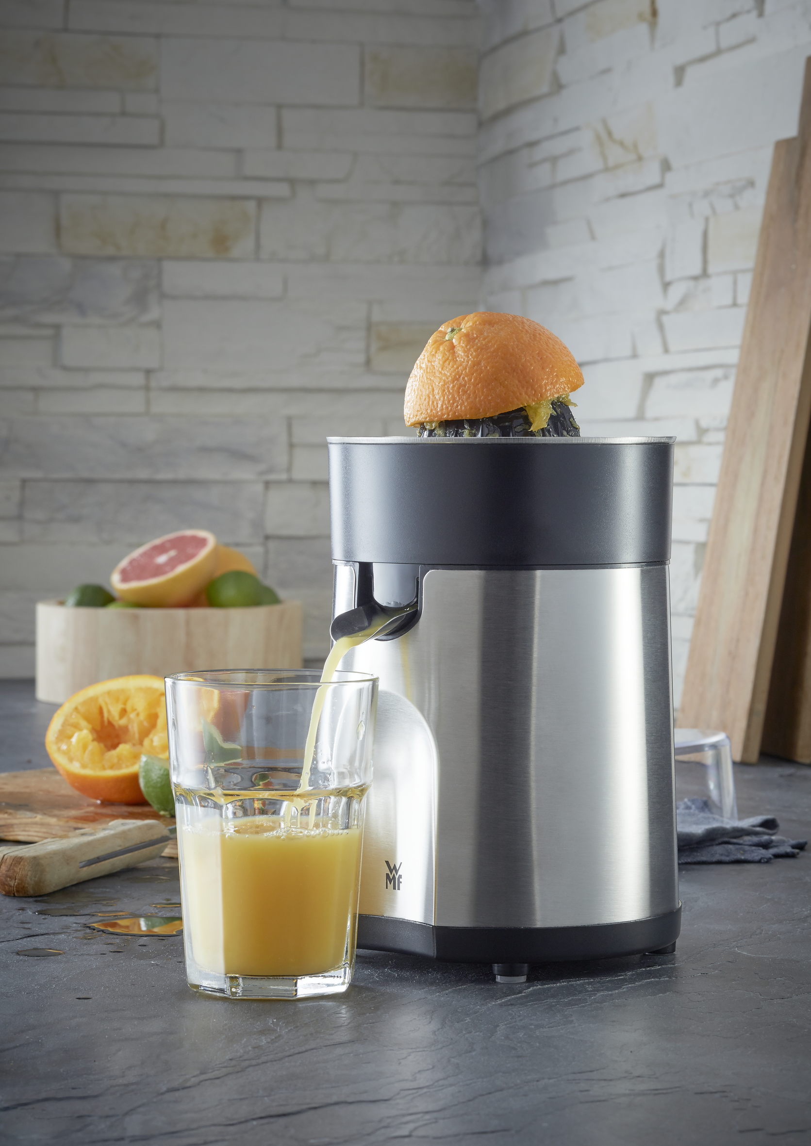 Exprimidor con mitad de naranja encima, vertiendo jugo en un vaso, otras frutas visibles.