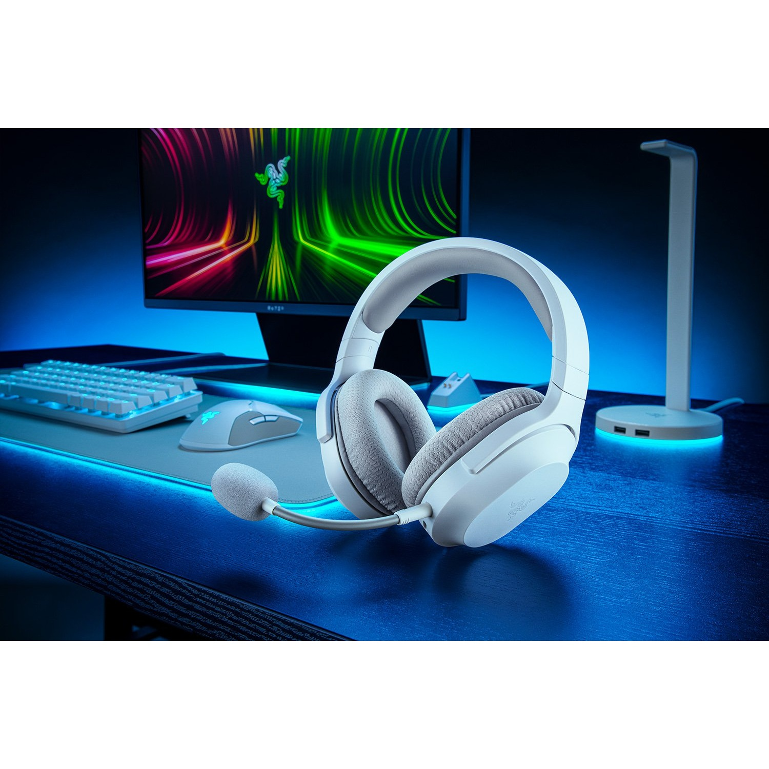 Auriculares gaming blancos con micrófono, teclado, ratón y monitor al fondo.