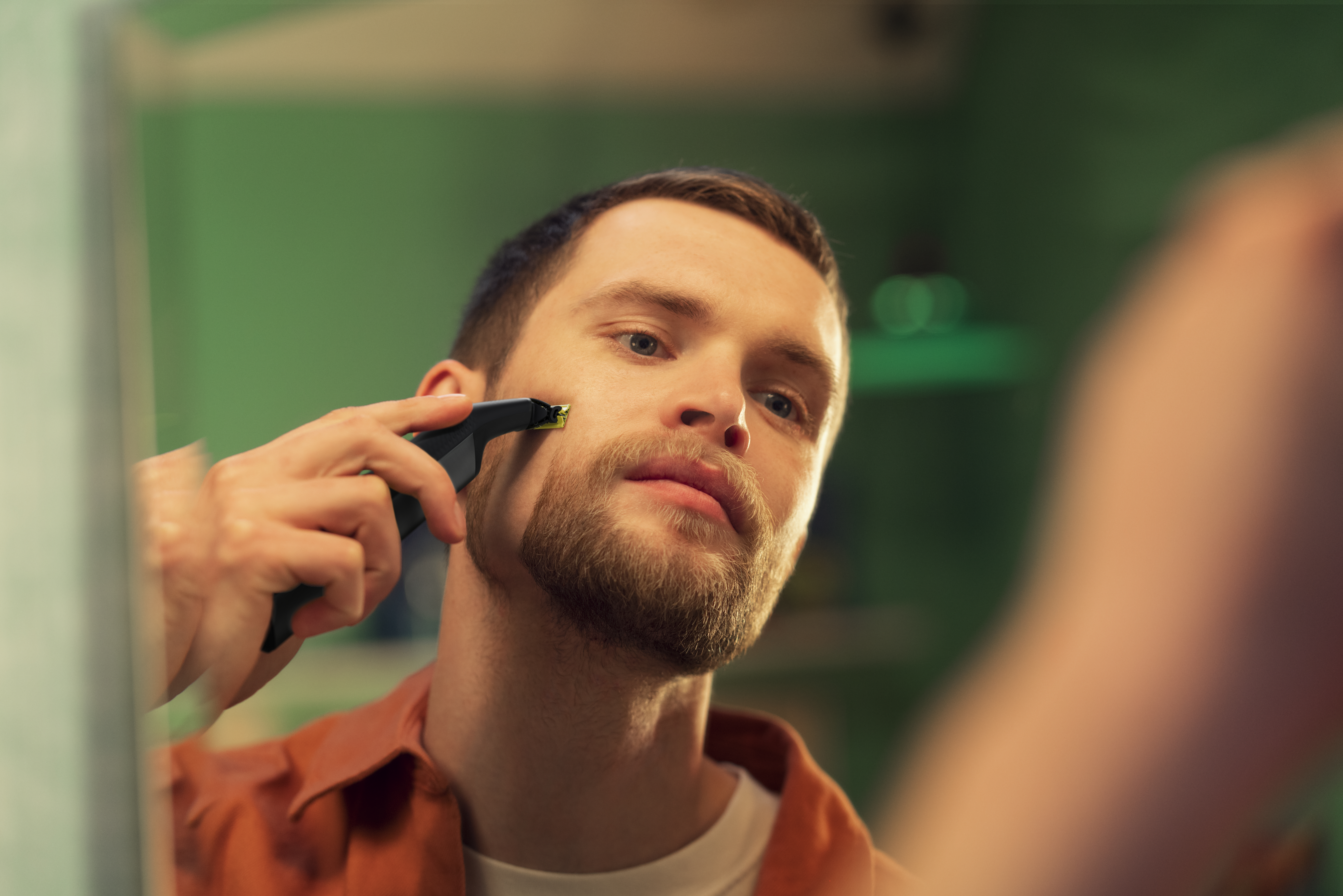 Ein Mann rasiert seinen Bart mit einem schwarzen Trimmer vor einem Spiegel. Grüner Hintergrund.