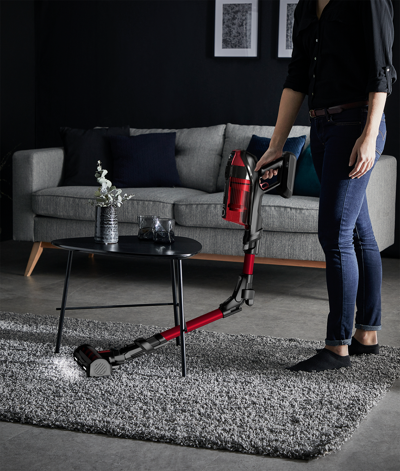 Woman using a red vacuum cleaner on a gray carpet. A sofa and table are in the background.