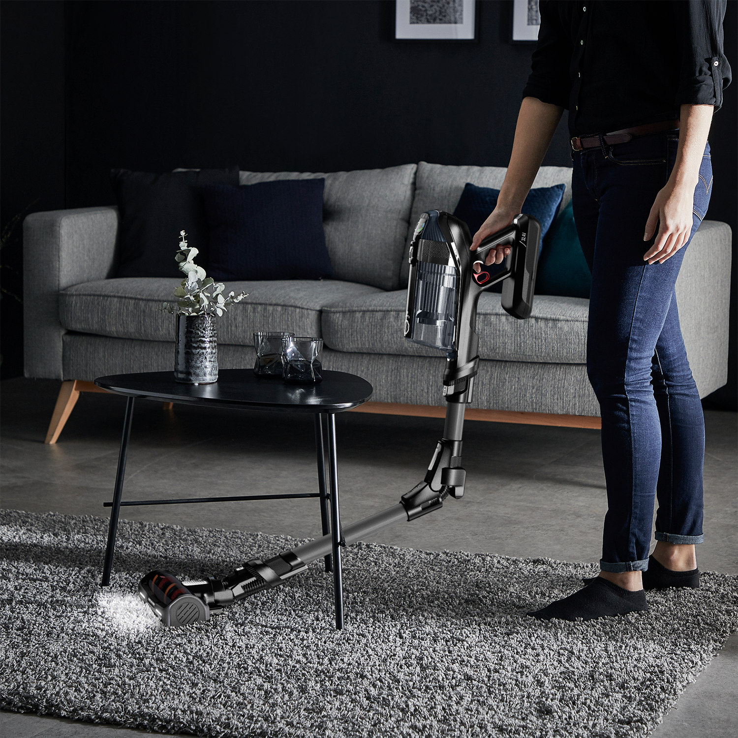 A woman using a black and red vacuum cleaner on a gray carpet, near a sofa and a table.