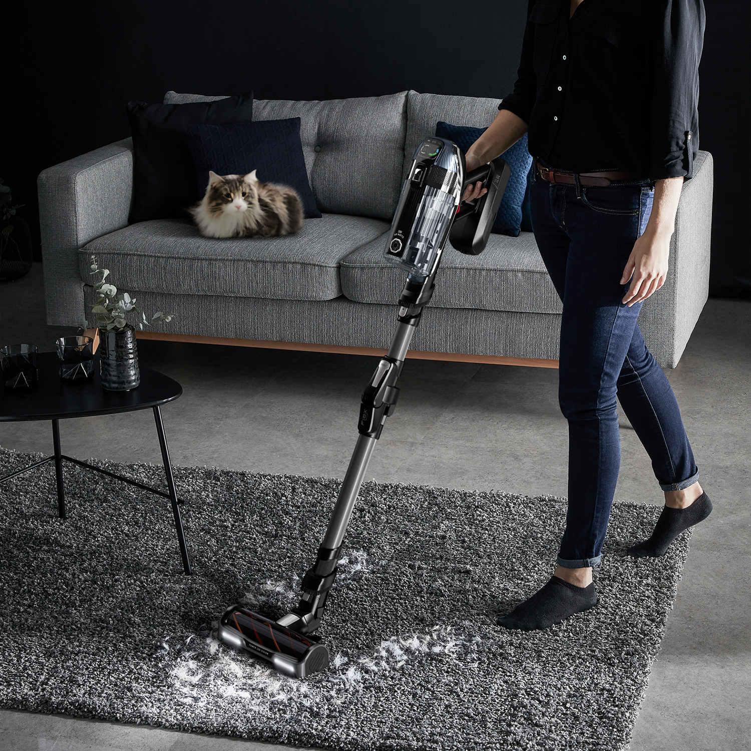 Woman vacuuming a rug, cat on couch in background. Sofa, table, and various home decor.