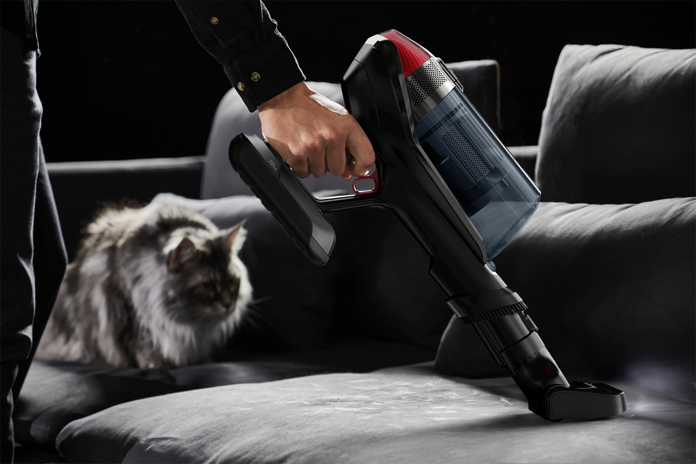 A person vacuuming a gray sofa with a black vacuum cleaner, a cat is in the background.