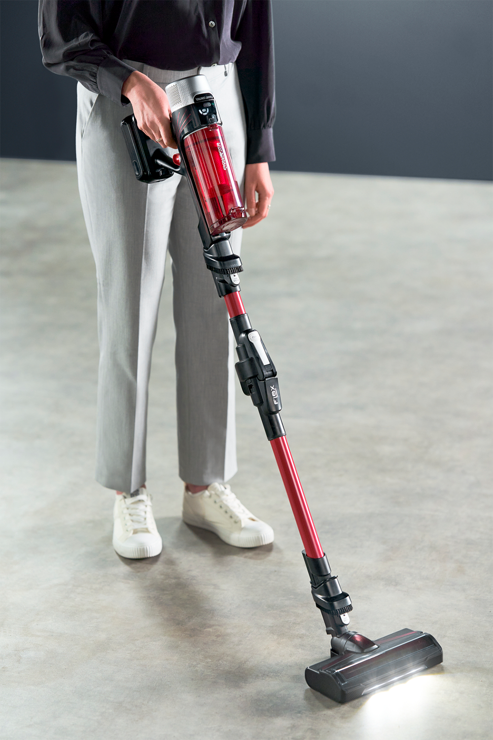 Een vrouw in een grijze broek en witte sneakers gebruikt een rood-zwarte snoerloze stofzuiger.