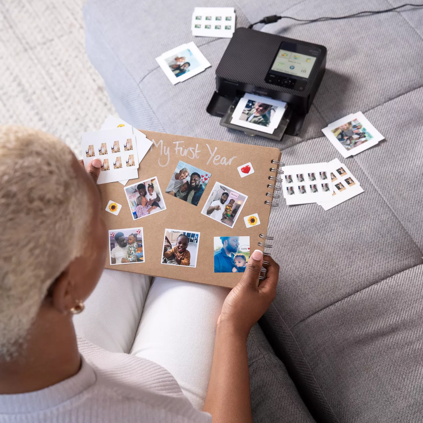 Una donna che tiene un album fotografico e delle foto. Una stampante sta stampando foto. L'album dice 'My First Year'.