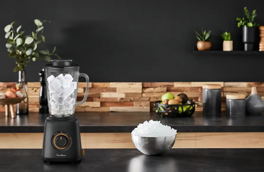 Blender with ice, next to a bowl of ice cubes, on a black countertop.