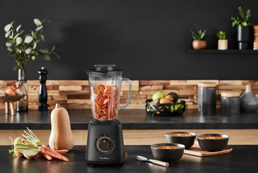 Blender with vegetables inside, next to bowls of soup on a black surface.