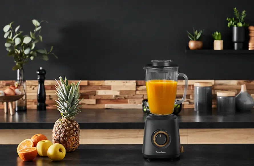 Blender with orange juice on a black countertop, with fruit and plants.
