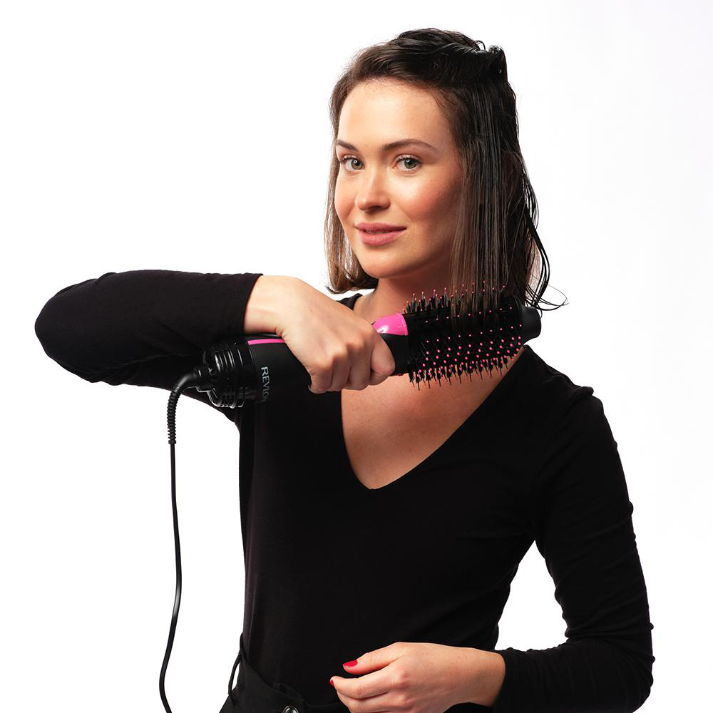 Frau benutzt eine pinke und schwarze Haartrocknerbürste, lächelt in die Kamera.