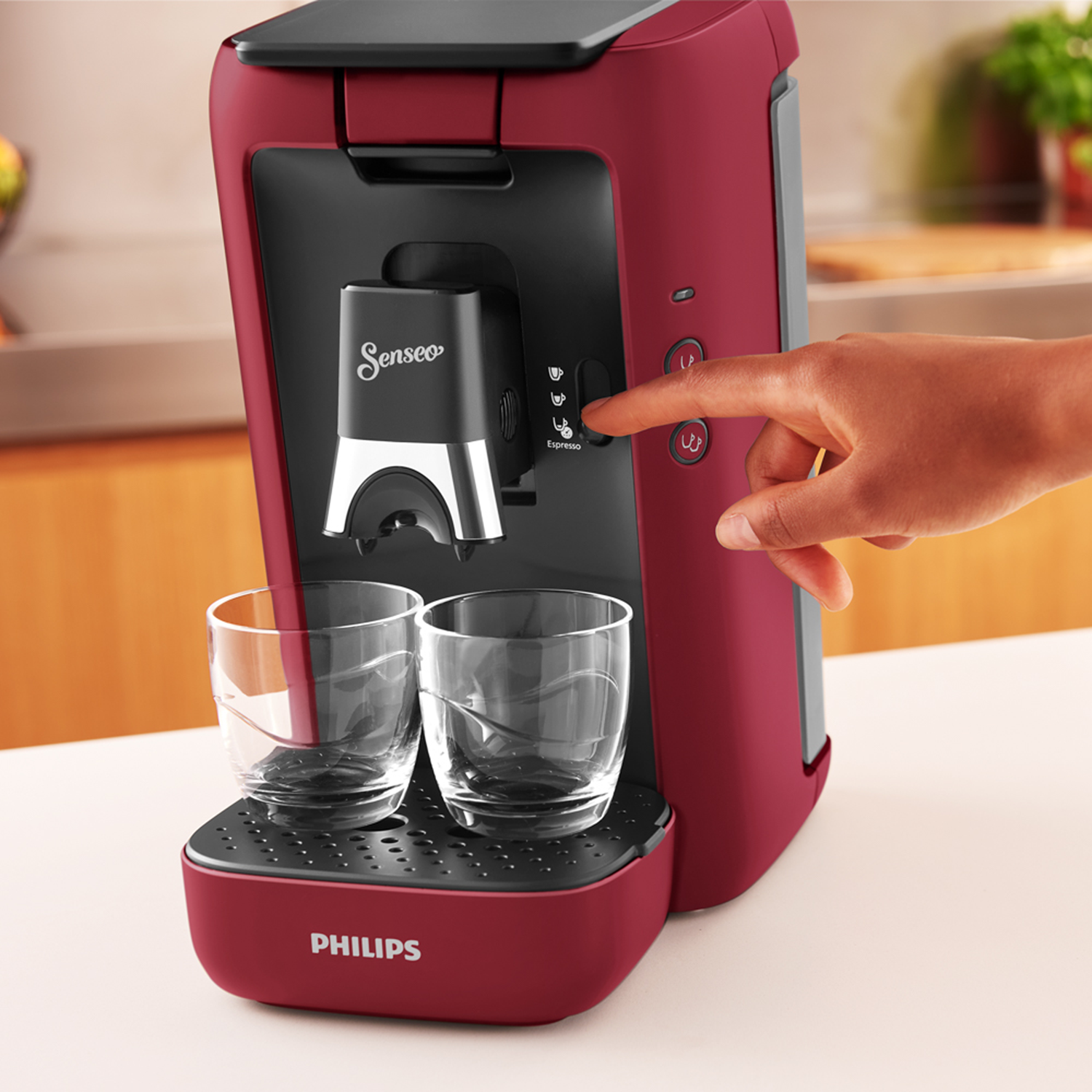 Rode Senseo koffiemachine met twee glazen en een hand die op een knop drukt. Witte achtergrond.