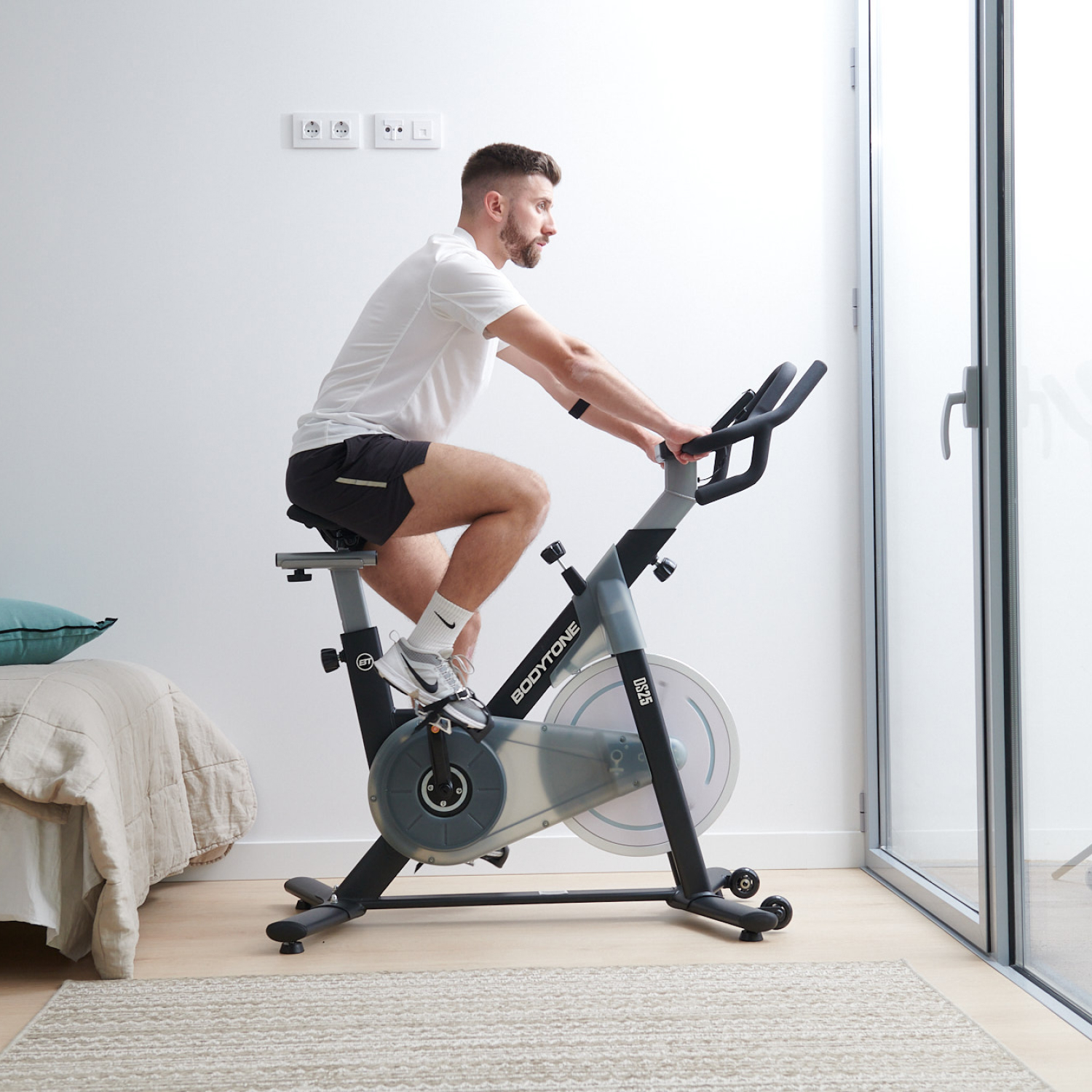 Un hombre montando una bicicleta estática en una habitación. Viste pantalones cortos y camiseta. Una ventana al fondo.