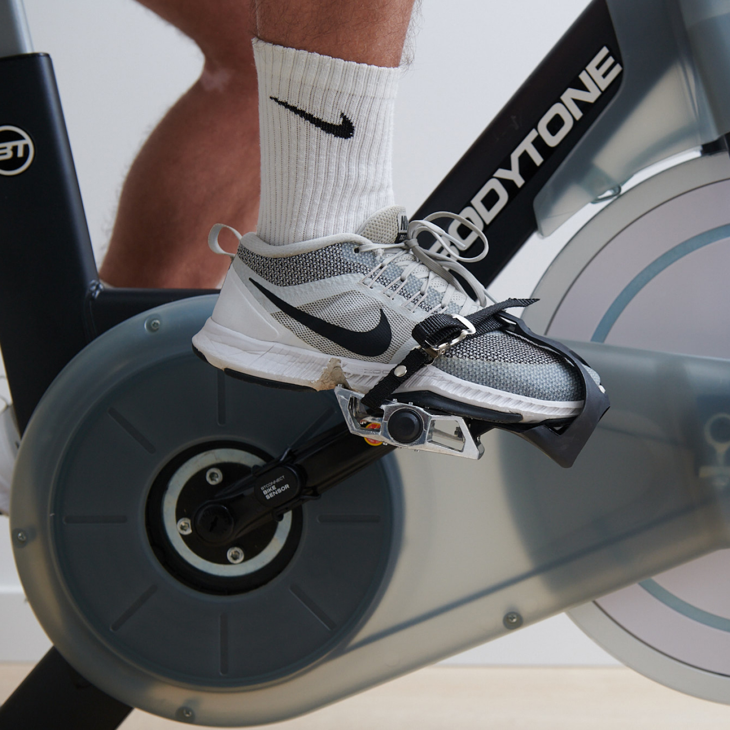 Pies de una persona con calcetines blancos y zapatillas en una bicicleta estática. Bicicleta negra y gris.