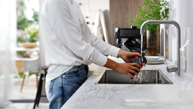 Person washes a black object under a kitchen faucet, water splashing. Kitchen in background.