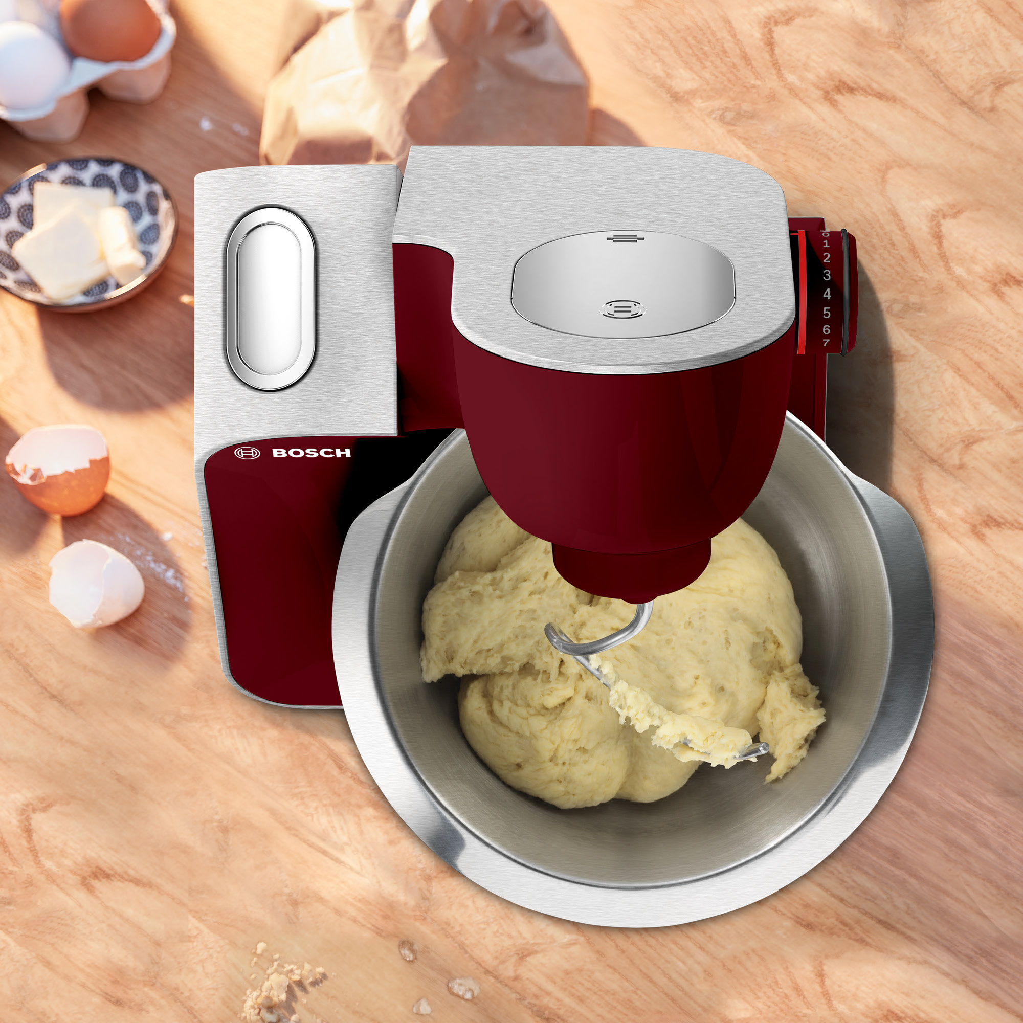 Une machine de cuisine Bosch rouge et argent avec un bol de pâte, ingrédients à proximité.