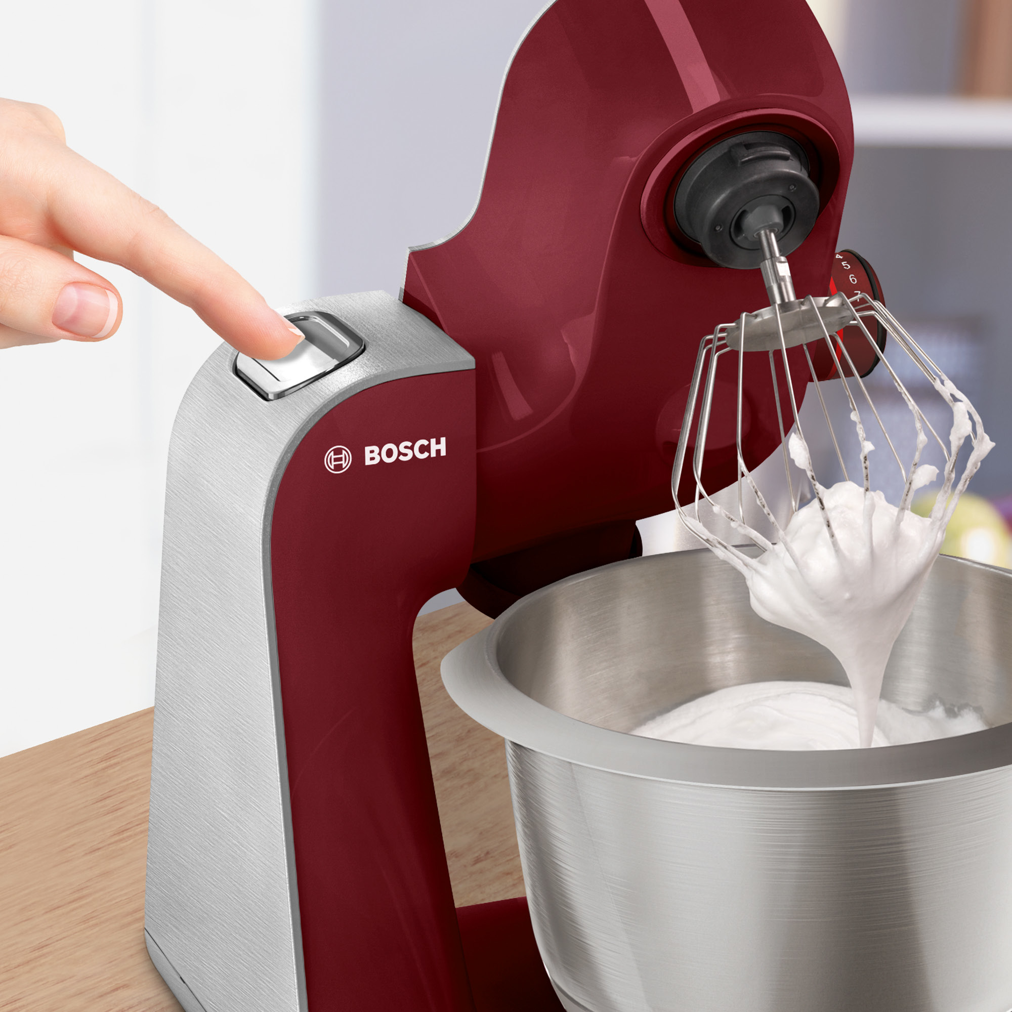 Une main appuie sur un bouton d'un robot de cuisine Bosch rouge et argent avec un fouet.