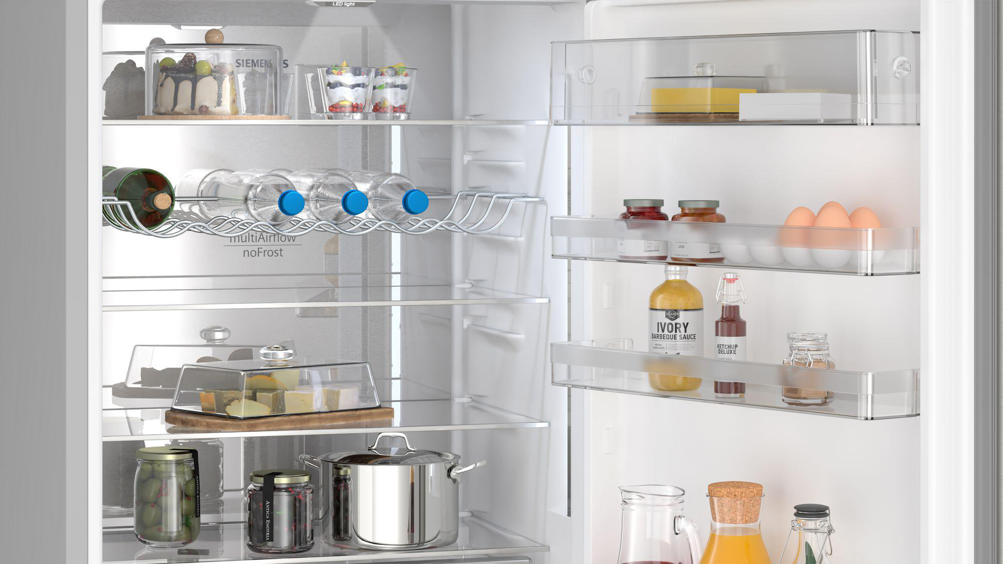 Offener Kühlschrank mit Regalen voller Lebensmittel, darunter Flaschen, Gläser und Behälter. Es gibt Eier und Kuchen.