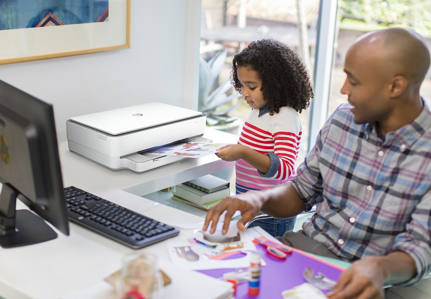 Een vader en dochter werken aan een bureau met een printer. Het meisje houdt een afdruk vast, met knutselspullen in de buurt.