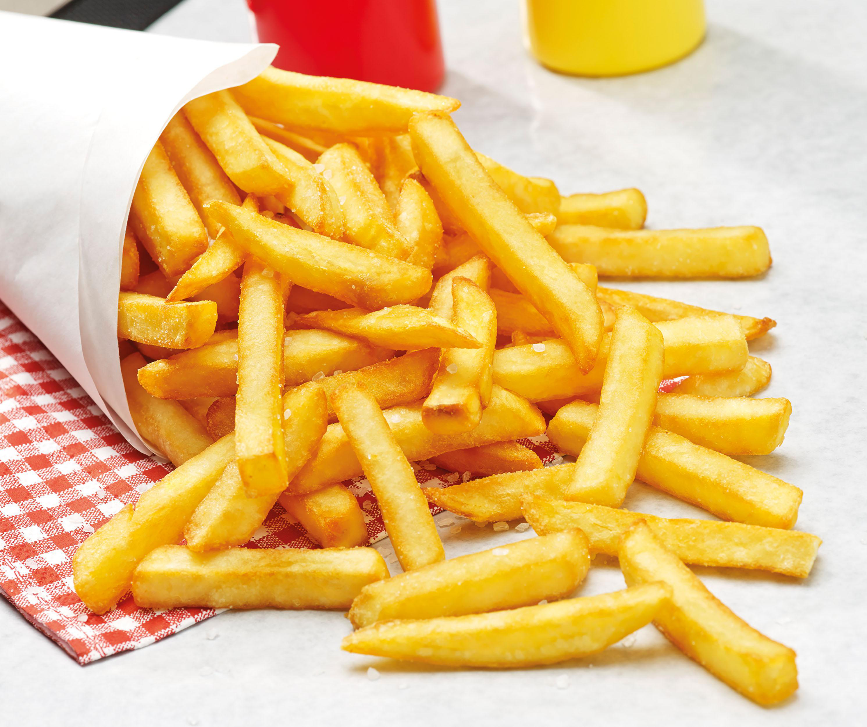 Pommes Frites, die aus einem Papierkegel auf eine Oberfläche fallen, rote und gelbe Gewürze.