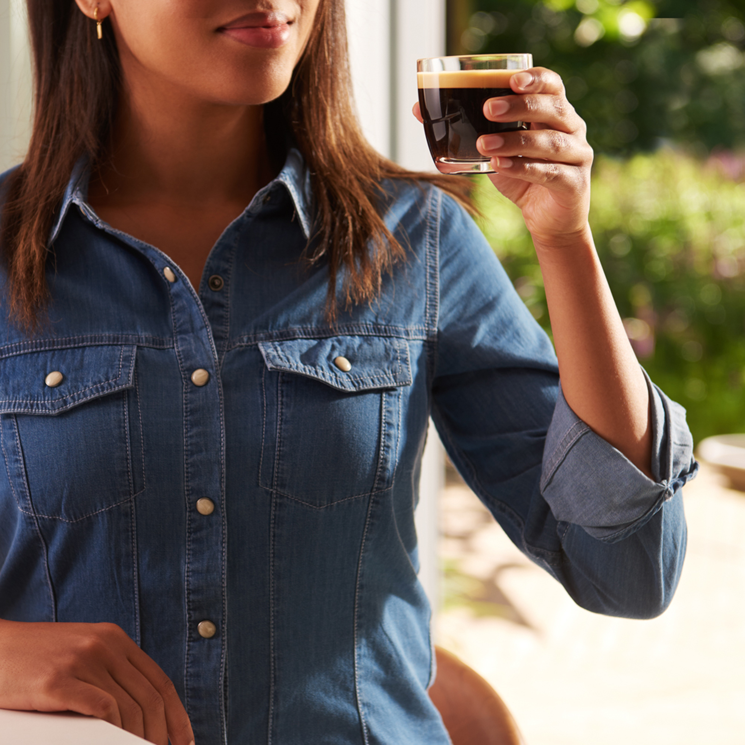 Woman holding a glass of dark coffee. She wears a denim shirt. Green background.