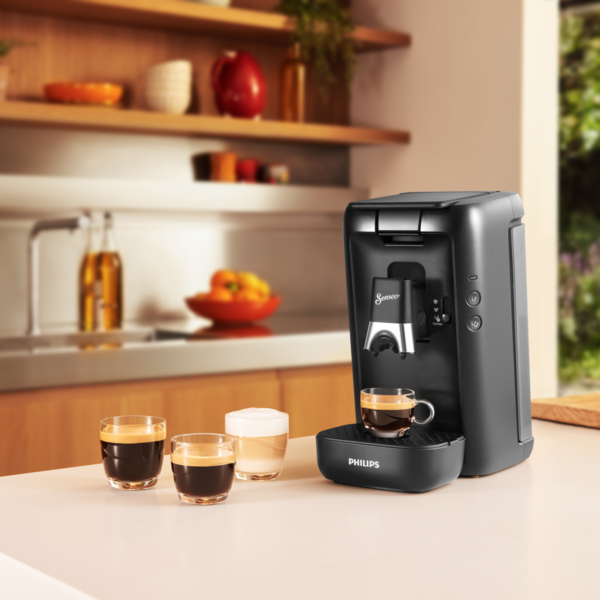 Black coffee machine with three cups of coffee, and a bowl of fruit on a white surface.