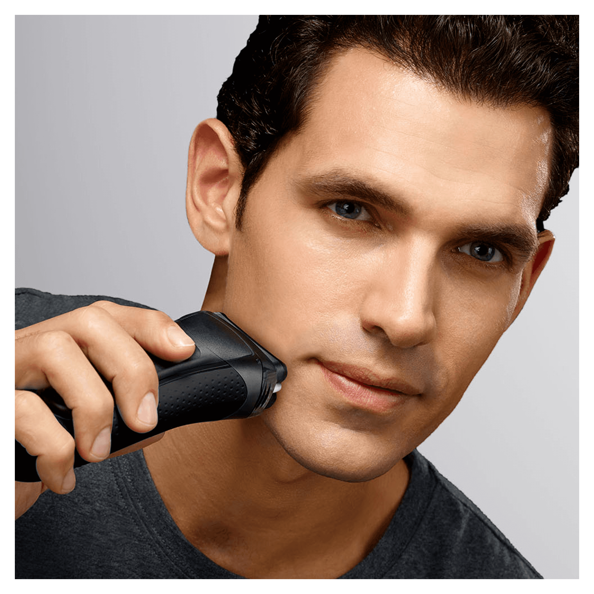 A man shaves his face with a black electric razor. He is looking at the camera.