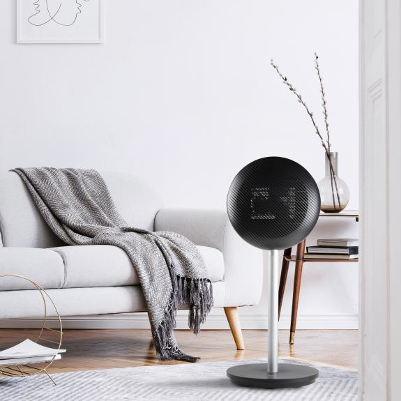 A modern black round speaker on a stand in a living room. A couch and decor are in the background.