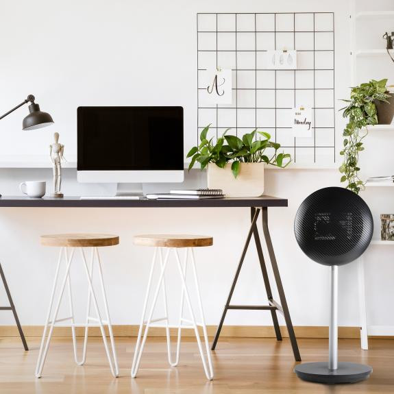 Modern office setting with desk, computer, stools, and a black fan. Plants and a grid board.