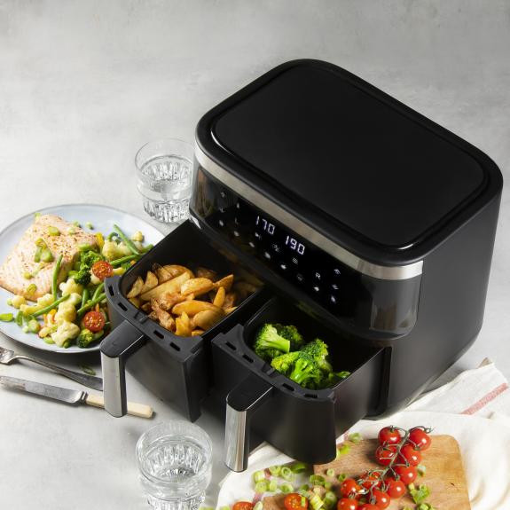 Black air fryer with two compartments, one with fries, the other with broccoli, with food on plates.