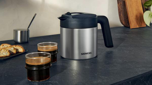 Eine silberne und schwarze Thermoskanne für Kaffee steht auf einer Theke neben Kaffeegläsern und Keksen.