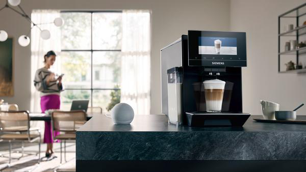 Eine Frau, die in einer Küche ein Telefon benutzt, mit einer Kaffeemaschine, die einen Latte zubereitet.
