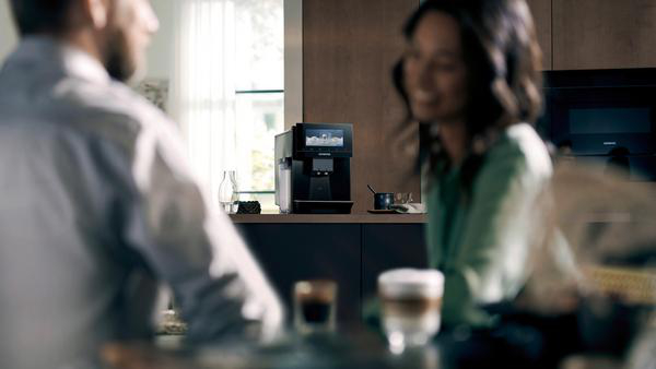 Personen in einer Küche, mit Kaffeemaschine und Kaffeetassen auf einem Tisch.