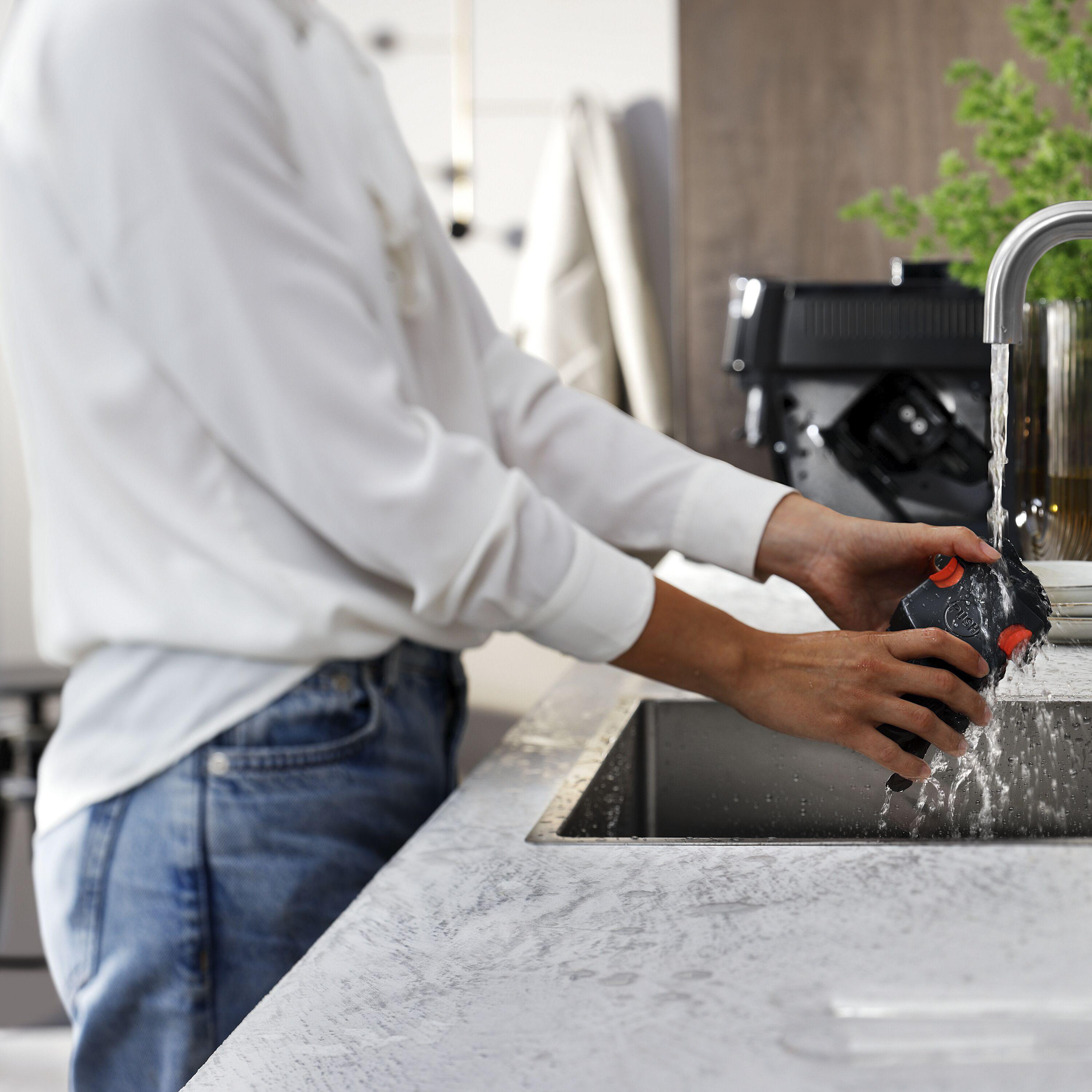 Eine Person wäscht ein schwarzes Objekt unter einem laufenden Wasserhahn in einem Edelstahlbecken. Der Wasserhahn ist verchromt.