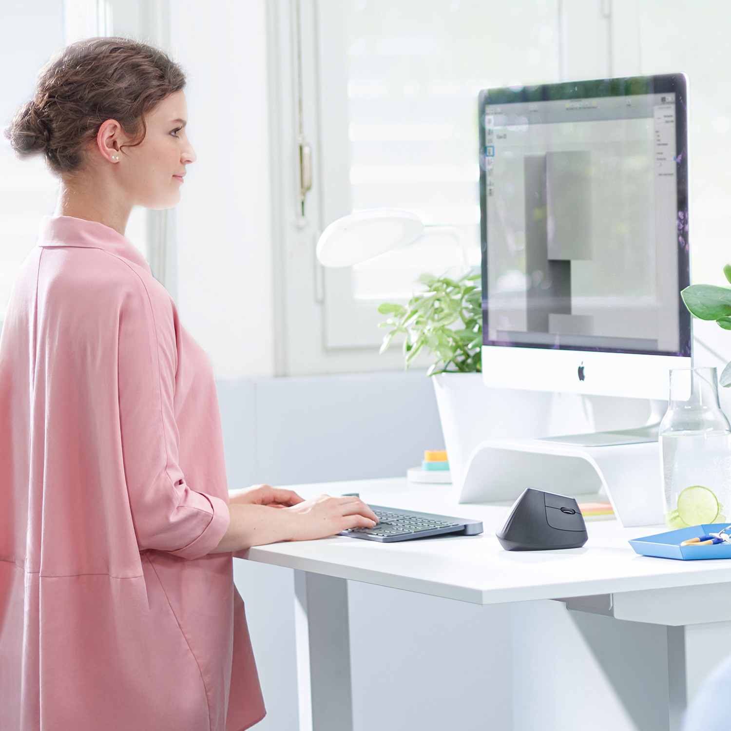 Mujer trabajando en un escritorio con computadora, teclado y ratón ergonómico.