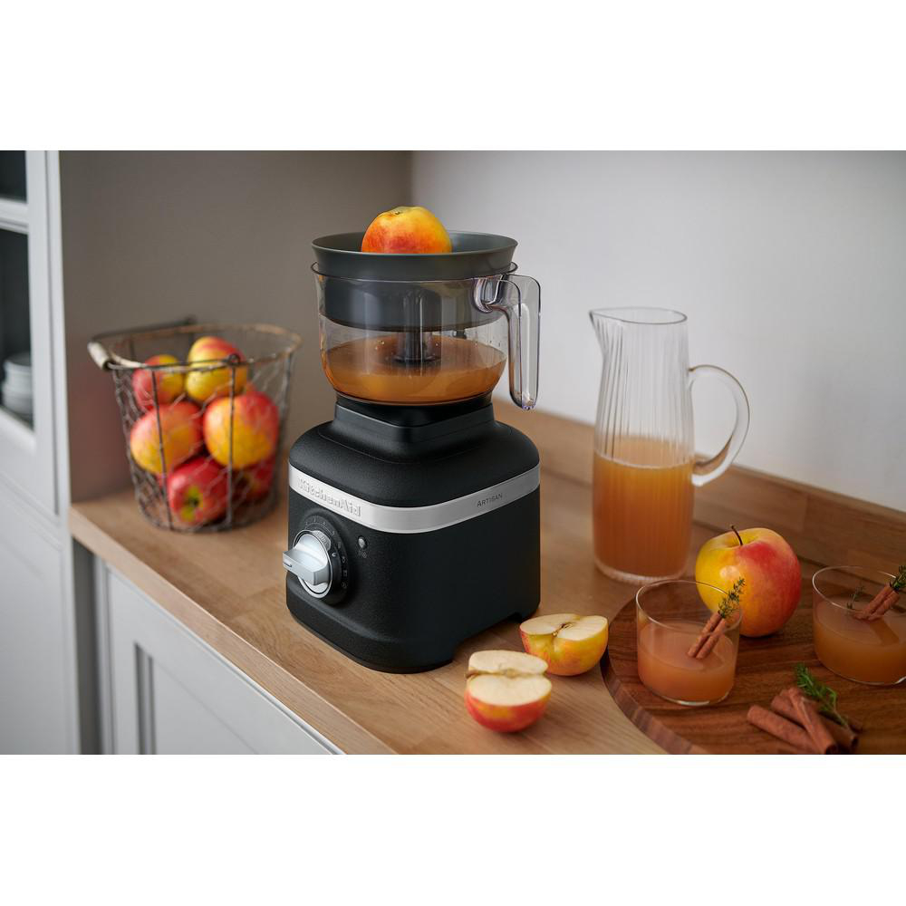 Schwarze KitchenAid Küchenmaschine mit Äpfeln, Saft und Zimtstangen auf einer Holzplatte.