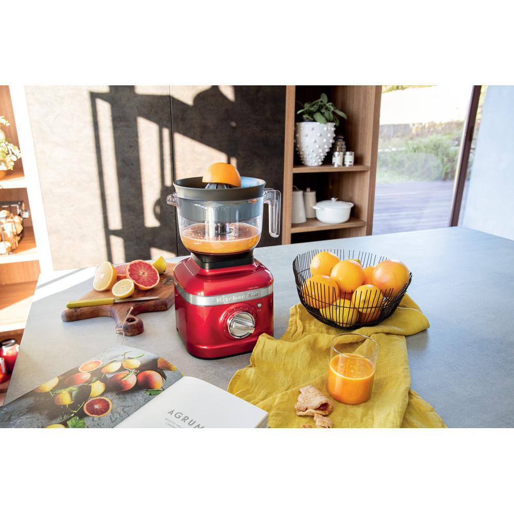 Ein roter KitchenAid-Entsafter mit Orangen und Zitronen auf einer Theke. Ein Glas Saft und eine Schüssel Orangen sind ebenfalls sichtbar.