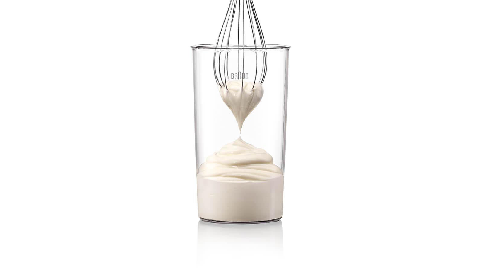 Ein klares Glas mit einem Schneebesen, Sahne wird in das Glas gegossen. Weißer Hintergrund.
