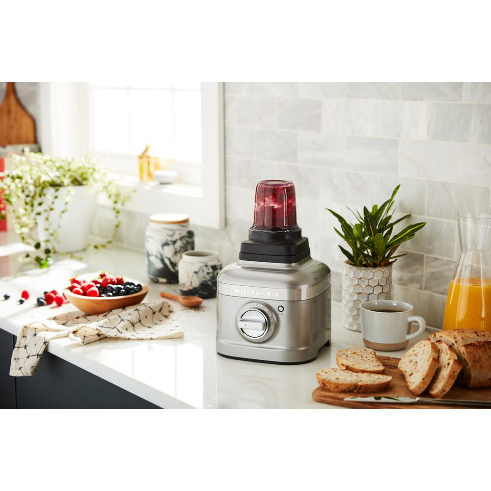 KitchenAid-Mixer mit rotem Smoothie. Beeren, Brot und Pflanze auf weißer Arbeitsplatte, Fenster im Hintergrund.