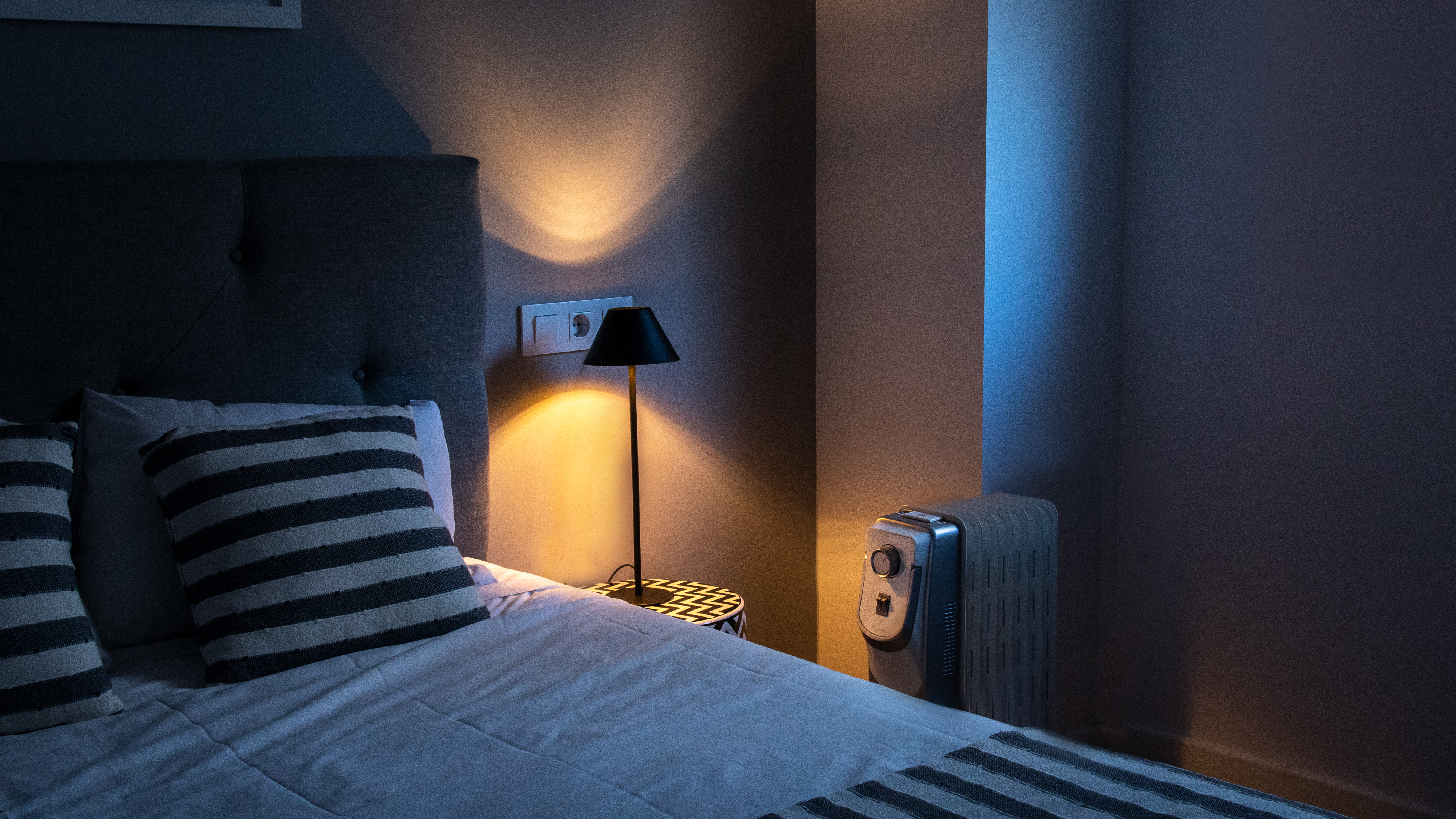 Una escena de dormitorio por la noche. Se ven una cama, una lámpara y un calefactor. La luz viene de la lámpara.