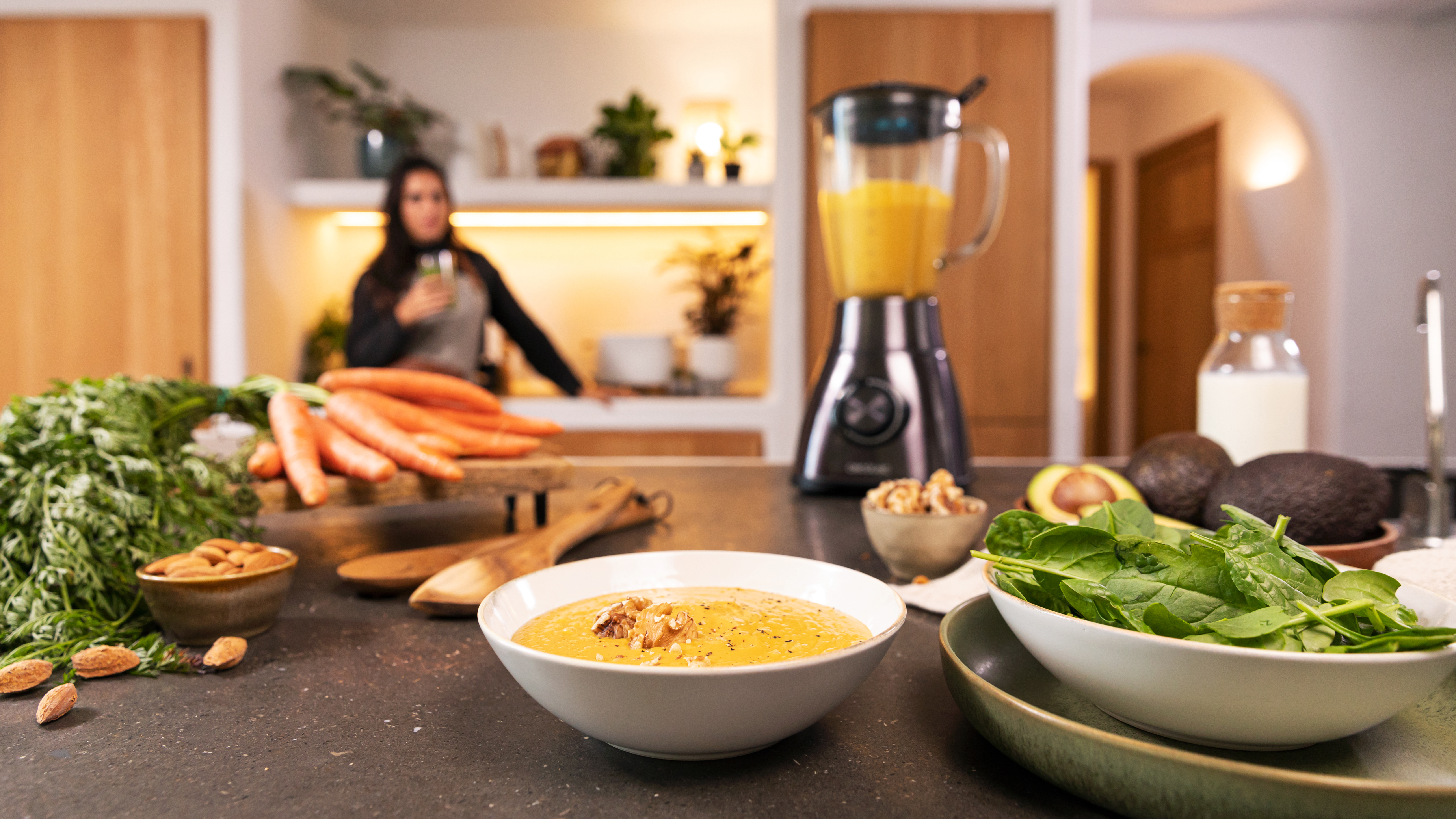 Escena de cocina con preparación de alimentos. Tazón de sopa, zanahorias, licuadora y mujer.