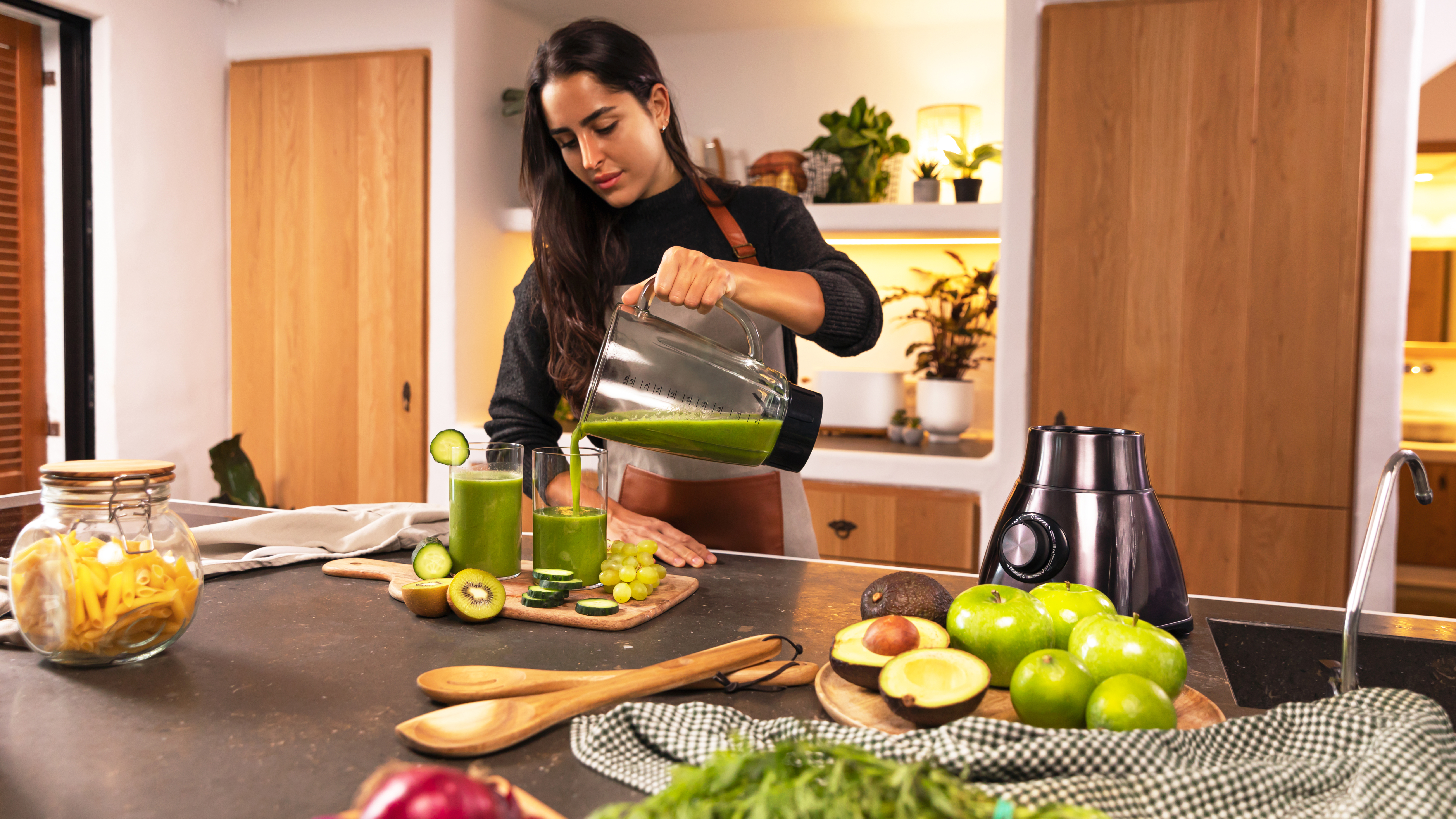 Mujer vertiendo jugo verde en vasos en una cocina, ingredientes alrededor.