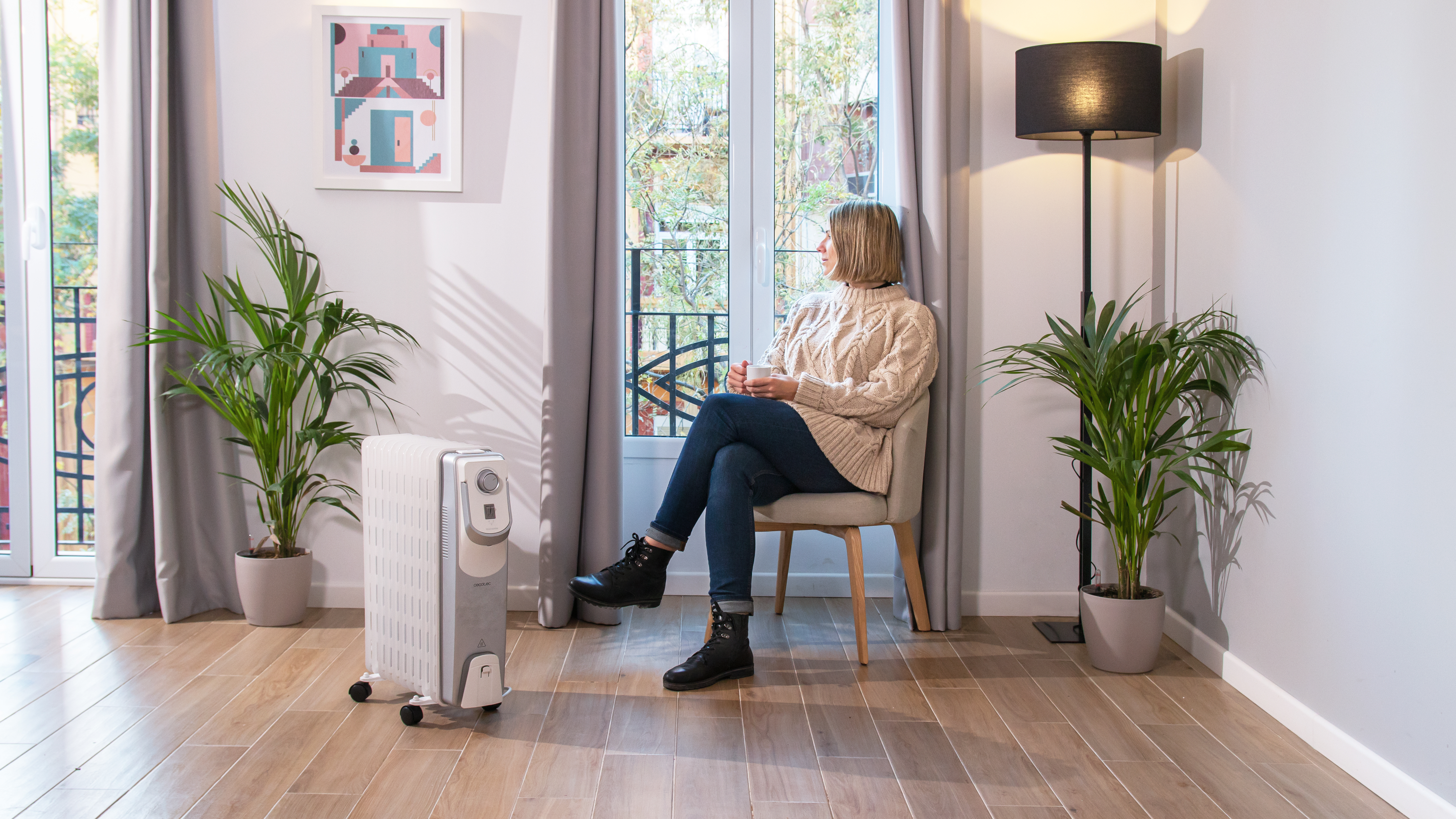 Mujer sentada en una habitación con un calefactor de aceite, plantas y ventana.